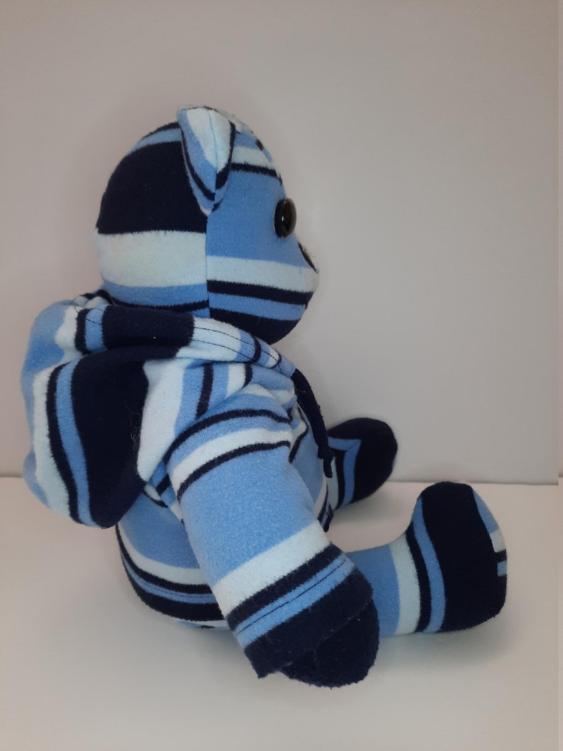 May include: A blue and navy striped plush teddy bear wearing a matching hooded sweatshirt. The bear is sitting with its legs crossed and has black button eyes.