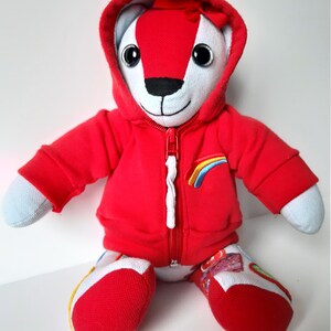 May include: A red hooded teddy bear with a white face and rainbow embroidery on the chest. The bear is sitting with its legs crossed and has a white zipper on its hoodie.