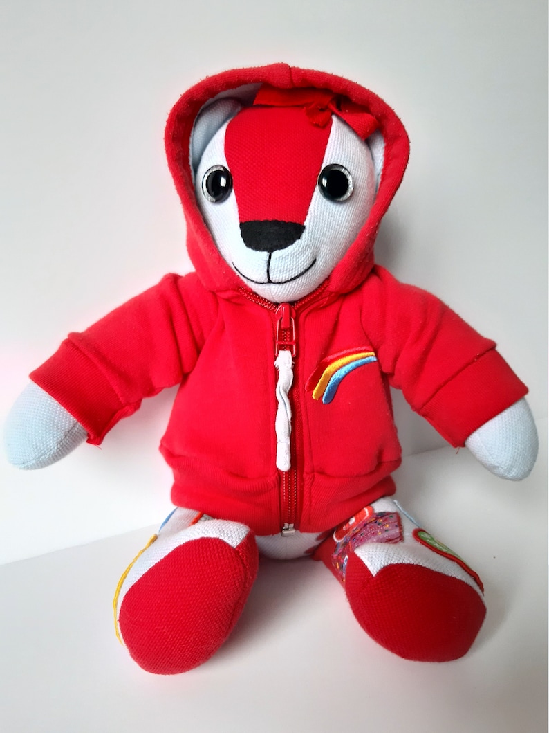 May include: A red hooded teddy bear with a white face and rainbow embroidery on the chest. The bear is sitting with its legs crossed and has a white zipper on its hoodie.