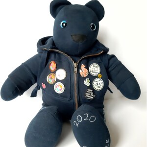 May include: A dark blue teddy bear wearing a hoodie with a zipper and several pins. The bear has blue eyes and a black nose. The bear's right foot has the text "2020 Graduate" embroidered on it.