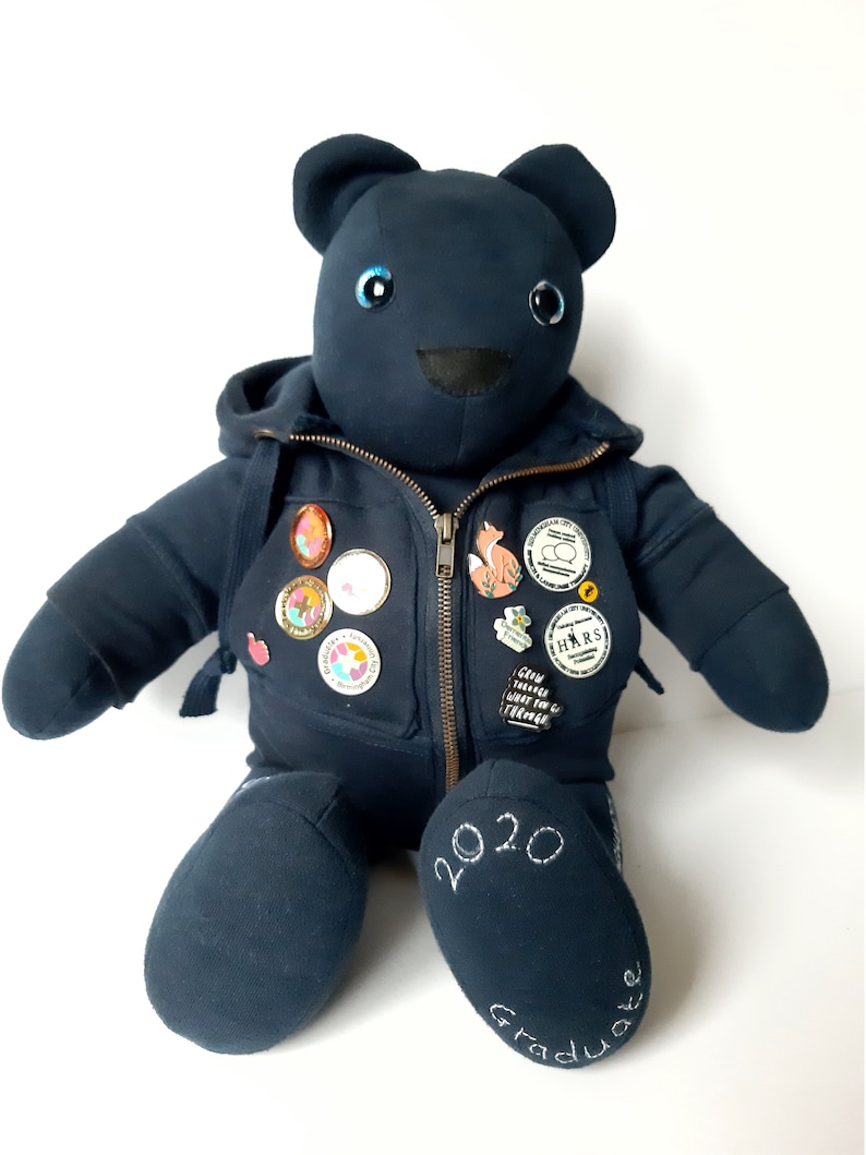 May include: A dark blue teddy bear wearing a hoodie with a zipper and several pins. The bear has blue eyes and a black nose. The bear's right foot has the text "2020 Graduate" embroidered on it.