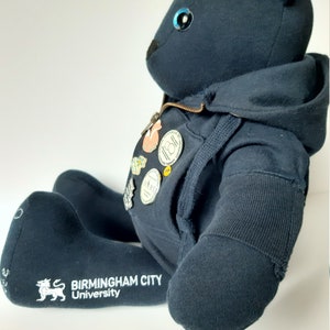 May include: Black teddy bear wearing a black hoodie with a zipper. The bear has blue eyes and is wearing several pins on its chest. The bear's foot has the text "Birmingham City University" embroidered on it.