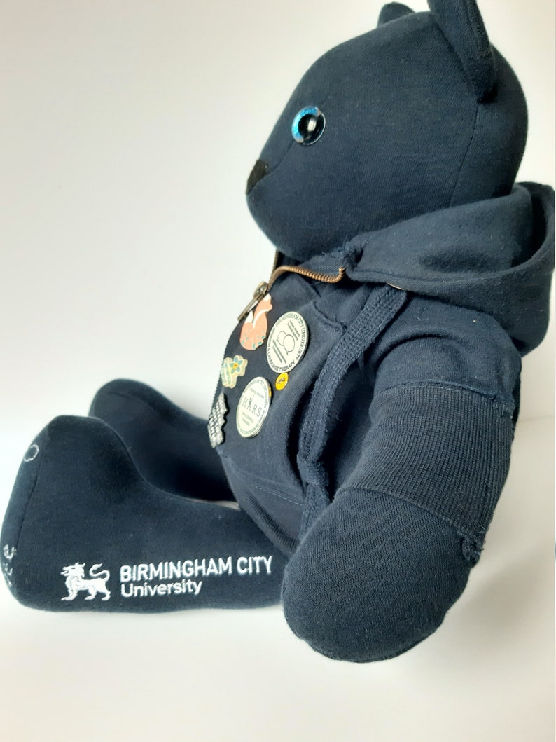 May include: Black teddy bear wearing a black hoodie with a zipper. The bear has blue eyes and is wearing several pins on its chest. The bear's foot has the text "Birmingham City University" embroidered on it.