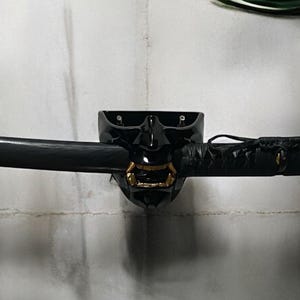 Può includere: Una spada katana nera con un supporto per maschera nero decorativo con accenti dorati. La spada è montata orizzontalmente contro un muro bianco strutturato, con vegetazione in primo piano e sullo sfondo.