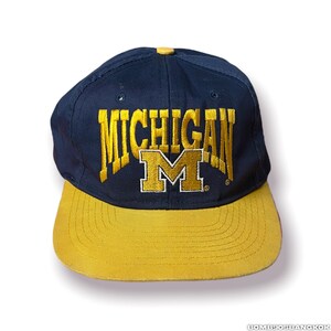 Puede incluir: Una gorra de béisbol azul marino y dorada con la palabra "MICHIGAN" y una gran "M" bordada en la parte delantera.
