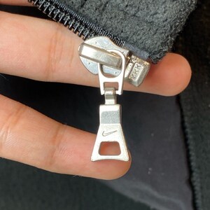 May include: Close-up of a silver zipper pull with a Nike swoosh logo. The zipper pull is attached to a black zipper with the text "YKK" on the slider.