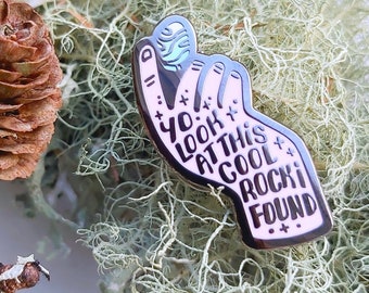 May include: A pink enamel pin with a hand holding a blue and white rock. The text on the pin reads "Yo! Look at this cool rock I found."