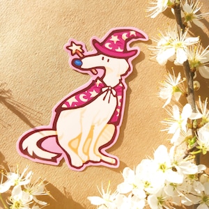 Könnte beinhalten: Ein Aufkleber mit einem Cartoon-Hund in einem Zaubererkostüm. Der Hund trägt einen rosa Zaubererhut mit weißen Sternen und Monden, einen passenden Umhang und einen sternförmigen Zauberstab. Der Aufkleber hat einen rosa Rand und einen beigen Hintergrund.