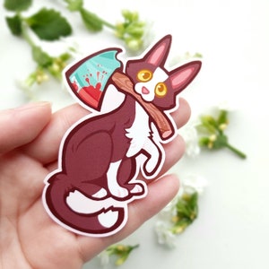 May include: A brown and white cat with yellow eyes holding a green and red axe. The cat is sitting on its hind legs and the axe is behind its back.