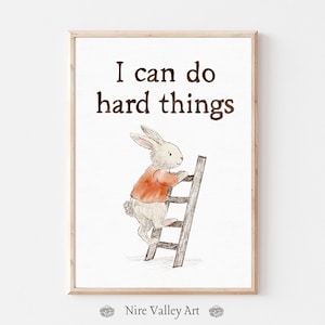 May include: A framed print featuring the text "I can do hard things" in brown. An illustration of a rabbit in an orange shirt climbing a wooden ladder is below the text. The print is in a light wood frame.