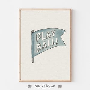 May include: A framed watercolor print featuring a blue flag with the words "PLAY BALL!" in white. The flag is attached to a gray pole. The artwork is set against a light beige background.