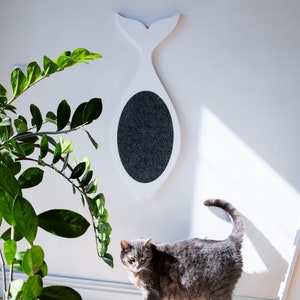 以下が含まれることがあります： 白い壁に取り付けられた、灰色の楕円形の引っ掻き面を備えた、クジラ型の白い猫用スクラッチャー。灰色のトラ猫がスクラッチャーの前の床に立っています。緑の植物が前景にあります。