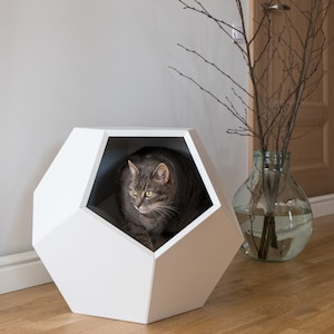 May include: A gray tabby cat sits inside a white geometric cat bed. The bed is shaped like a dodecahedron and has a large opening on one side. The cat is looking out of the opening.