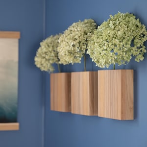 Puede incluir: Tres jarrones de pared de madera, cada uno con un racimo de flores verde claro, están montados en una pared azul. Una impresión de paisaje enmarcada cuelga en la pared adyacente.
