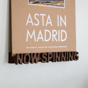 May include: A dark brown wooden display stand featuring the cut-out text NOW SPINNING, holding a tan rectangular print with the white text ASTA IN MADRID and a small landscape image at the top, mounted against a plain white wall surface.