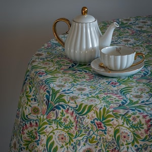 Puede incluir: Un juego de té de porcelana blanca con detalles dorados descansa sobre un mantel redondo. La tetera y la taza tienen un diseño acanalado. El mantel presenta un estampado floral con flores rosas, verdes y azules.