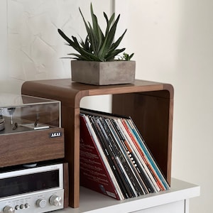 May include: A wooden record shelf with a built-in stand for a turntable. The shelf is filled with vinyl records and a small potted succulent plant sits on top. The shelf is labeled "AKAI".