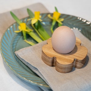 May include: A brown egg sits in a wooden egg cup shaped like a flower. The egg cup is on a beige linen napkin, which is on top of a blue and green ceramic plate.  There are yellow daffodils in the background.