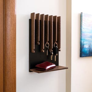 Op de afbeelding: Een houten sleutelhouder met een zwarte achterkant en een klein plankje. De sleutelhouder heeft verschillende verticale houten latten om sleutels aan op te hangen. Een rode portemonnee ligt op de plank. De sleutelhouder is aan een witte muur bevestigd.