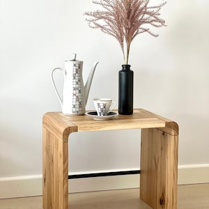 May include: A wooden side table with a curved design, featuring a white teapot, a small teacup and saucer, and a vase with dried pink pampas grass.