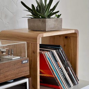 May include: A light-toned wooden record storage cube with rounded edges, holding a collection of vinyl records. A rectangular concrete planter with a green succulent sits on top. To the left, a wooden Akai turntable is partially visible.