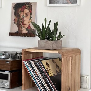 May include: A wooden record stand with a curved design holds a stack of vinyl records. A potted succulent plant sits on top of the stand. The stand is on a white shelf with a record player and a light switch.