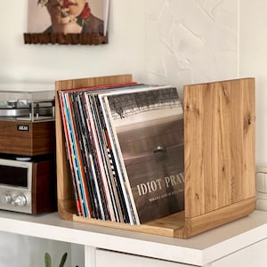 May include: A wooden record storage shelf with a stack of vinyl records. The top record has the title "Idiot Pray" by Nick Cave and the Bad Seeds.