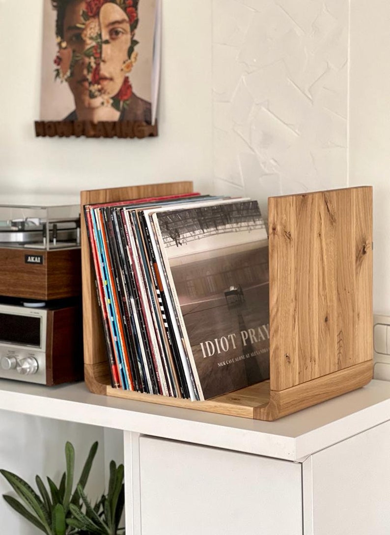 May include: A wooden record storage shelf with a stack of vinyl records. The top record has the title "Idiot Pray" by Nick Cave and the Bad Seeds.