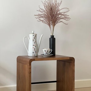May include: A wooden side table with a curved top, featuring a white teapot, a white teacup and saucer, and a vase with dried pink pampas grass.