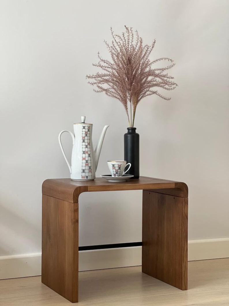 May include: A wooden side table with a curved top, featuring a white teapot, a white teacup and saucer, and a vase with dried pink pampas grass.