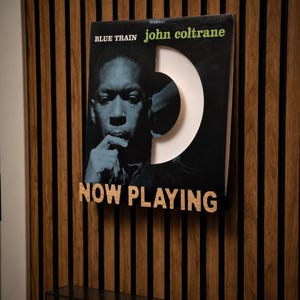 May include: A framed John Coltrane "Blue Train" album cover with a unique cutout design. The album art features a blue-toned portrait of Coltrane. Below the album is a wooden sign that reads "NOW PLAYING". The background is a wooden wall.