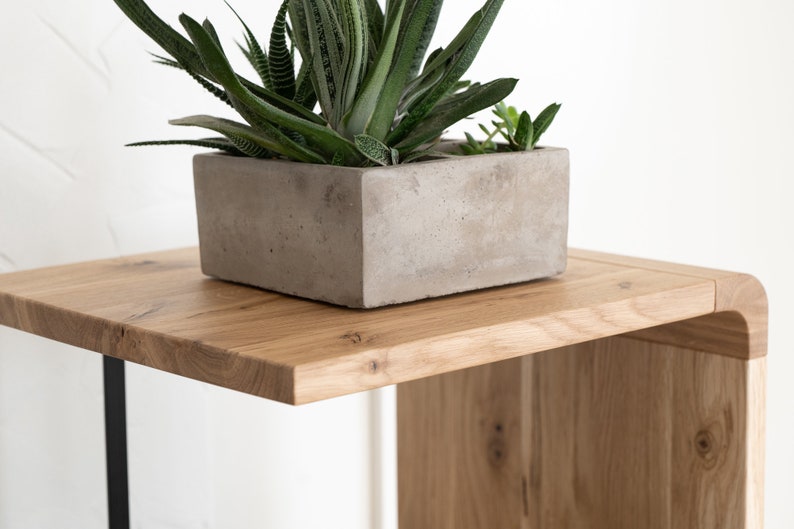May include: A rectangular concrete planter containing a green succulent plant with pointed leaves, positioned on a light brown wooden side table with a smooth surface and a curved edge, featuring a visible wood grain and a thin black metal leg.
