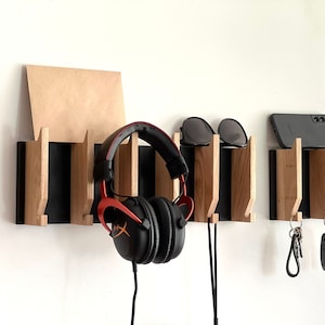 Op de afbeelding: Een houten wandorganizer met zwarte accenten. Het heeft drie haken om koptelefoons, zonnebrillen en sleutels op te hangen. De organizer is gemaakt van lichtbruin hout en heeft een modern ontwerp.