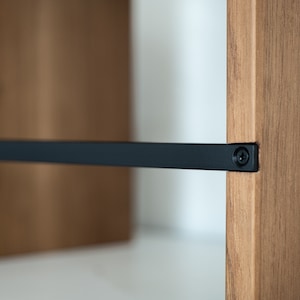 May include: Close-up of a wooden shelf with a black metal support bar attached with a screw. The shelf is made of natural wood with a light brown finish.