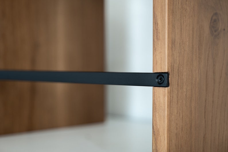 May include: Close-up of a wooden shelf with a black metal support bar attached with a screw. The shelf is made of natural wood with a light brown finish.
