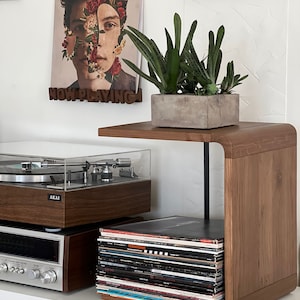 May include: A wooden record player stand with a shelf for vinyl records. The stand has a unique curved design and a black metal support. A stack of vinyl records is on the shelf, and a record player is on the top shelf. A potted plant sits on the top shelf of the stand. The stand is against a white wall with a framed picture and a poster with the words "NOW PLAYING" above it.