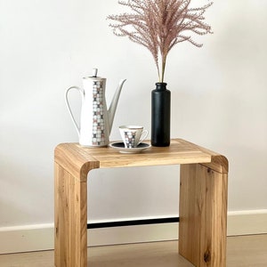 May include: A light-coloured wooden side table with a curved design. On top sits a white coffee pot with a geometric grid pattern, a matching teacup and saucer, and a matte black bottle containing a tall, feathery, light brown dried plant.