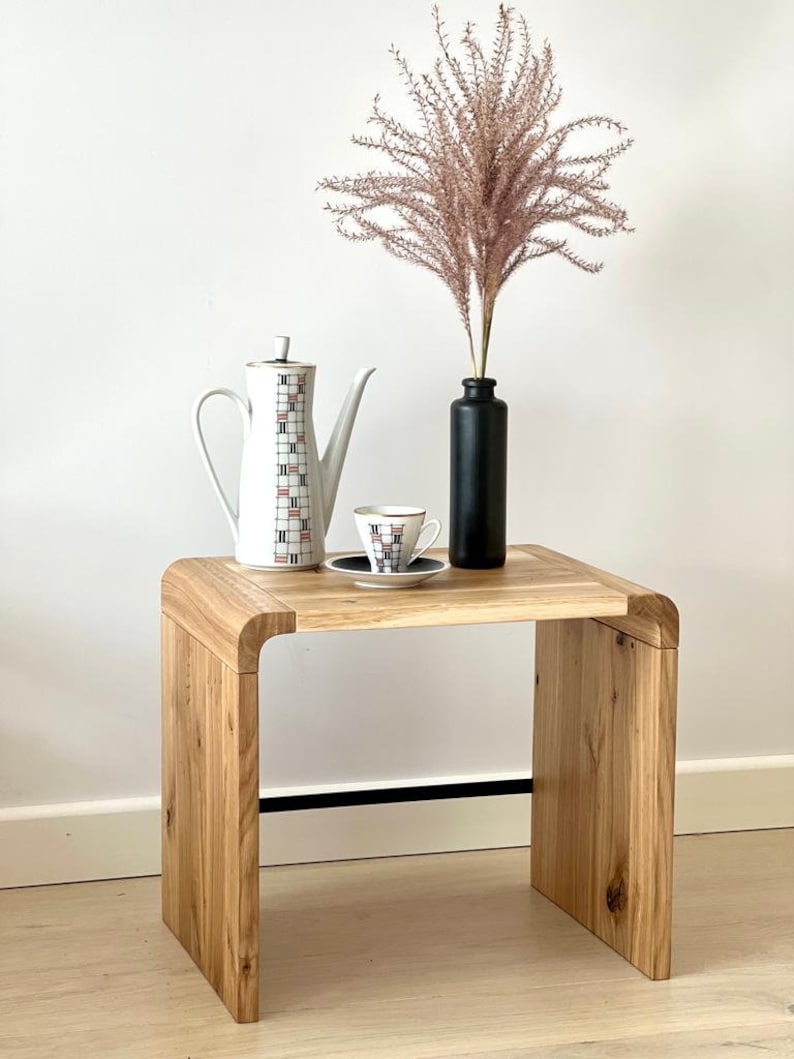 May include: A light-coloured wooden side table with a curved design. On top sits a white coffee pot with a geometric grid pattern, a matching teacup and saucer, and a matte black bottle containing a tall, feathery, light brown dried plant.