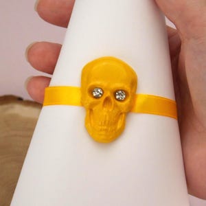 Può includere: Un oggetto a forma di cono bianco è adornato con un teschio giallo e un nastro giallo abbinato. Il teschio ha occhi di cristallo. L'oggetto è tenuto in mano, suggerendo che si tratti di un oggetto decorativo, forse per Halloween o un evento a tema.