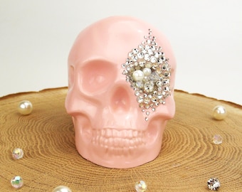 May include: A pink ceramic skull with a rhinestone and pearl eye patch. The skull is sitting on a wooden surface.