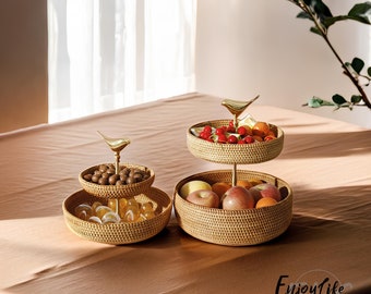 May include: Two-tiered woven wicker fruit basket with gold bird accents. The top tier holds red cherries, the middle tier holds brown nuts, and the bottom tier holds clear jelly candies. The basket is on a beige tablecloth.