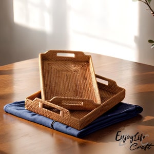 Puede incluir: Dos bandejas de mimbre trenzado con asas, apiladas una encima de la otra. Las bandejas son de color marrón claro y están hechas de materiales naturales. Las bandejas están sobre un paño azul sobre una superficie de madera.