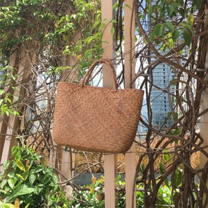 Pu&ograve; includere: Una borsa tote in vimini marrone con una trama in paglia naturale. La borsa &egrave; appesa a una recinzione di legno.