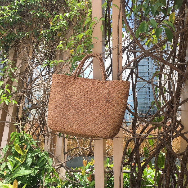 Pu&ograve; includere: Una borsa tote in vimini marrone con una trama in paglia naturale. La borsa &egrave; appesa a una recinzione di legno.