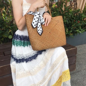 Pu&ograve; includere: Una donna tiene in mano una borsa di paglia intrecciata con un nastro a pois bianchi e neri. La borsa ha una trama intrecciata ed &egrave; di colore marrone chiaro. La donna indossa un vestito bianco con bordi in pizzo e una striscia verde e blu.
