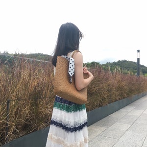 Pu&ograve; includere: Una donna indossa un vestito bianco e verde con una borsa tote in vimini marrone con una sciarpa a pois bianchi e neri. La borsa &egrave; appesa alla sua spalla.
