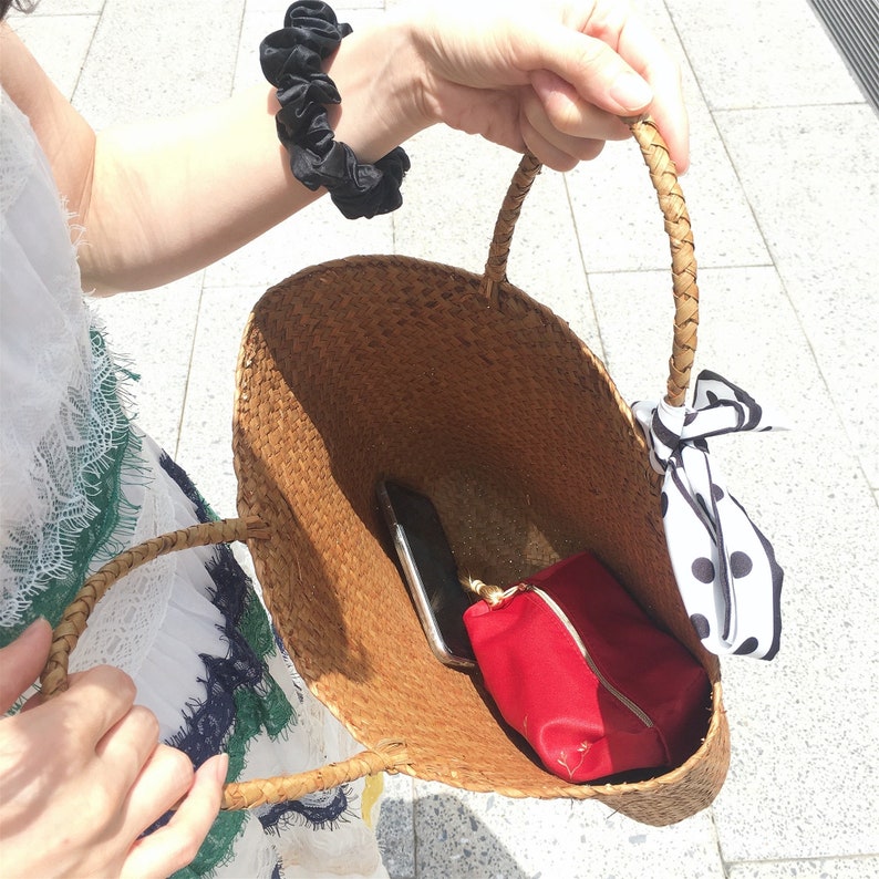 Pu&ograve; includere: Una grande borsa tote in vimini marrone con un fiocco in tessuto a pois bianchi e neri. La borsa &egrave; aperta e contiene una pochette con cerniera rossa e un telefono cellulare nero.