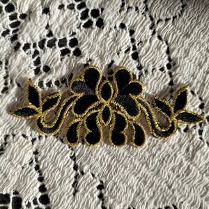 May include: Black floral applique with gold trim. The applique features a symmetrical design of stylized flowers and leaves. The gold trim outlines the black fabric, creating a decorative embellishment.