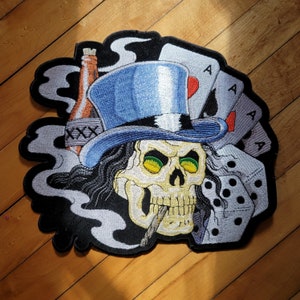 May include: Embroidered patch featuring a skull wearing a blue top hat, smoking a cigarette, and surrounded by playing cards, dice, and a bottle. The skull has green eyes and long black hair. The patch has a black border and a smoky background.