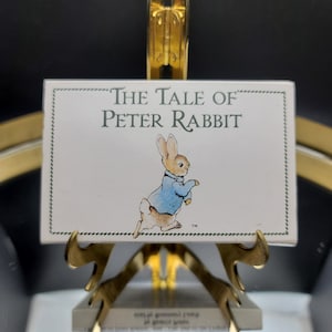 May include: A rectangular sign featuring the title "The Tale of Peter Rabbit" with an illustration of Peter Rabbit in a blue jacket. The sign is displayed on a gold-colored easel, with a black background.